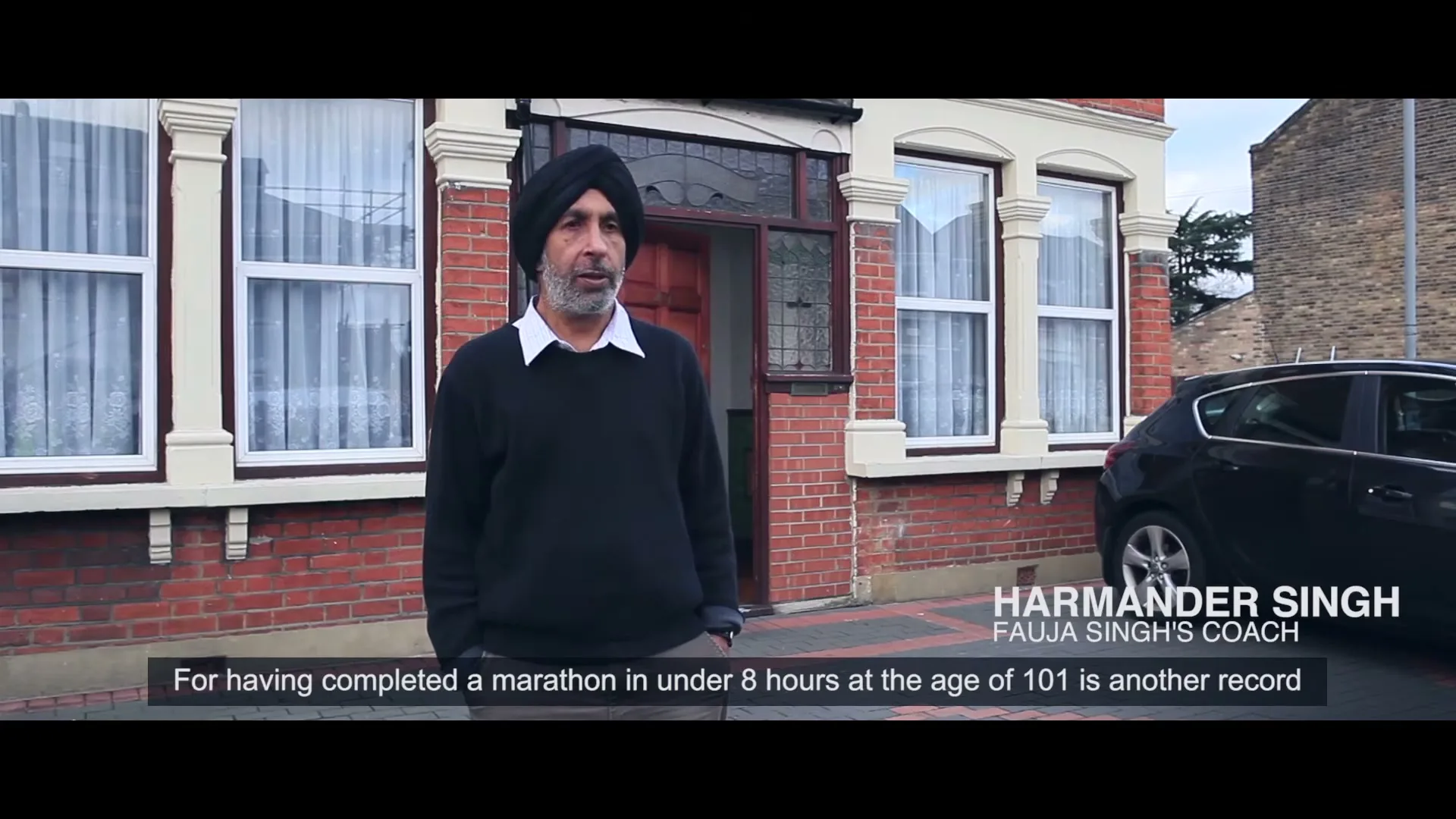 Harmander Singh, Fauja Singh's coach, stands outside a brick house with white windows and a black car parked nearby, speaking to the camera.