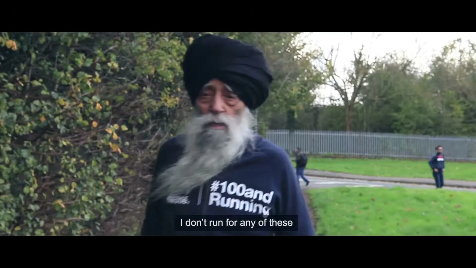 An elderly Sikh man with a long white beard and black turban runs outdoors, wearing a blue sweatshirt with "#100andRunning" printed on it.