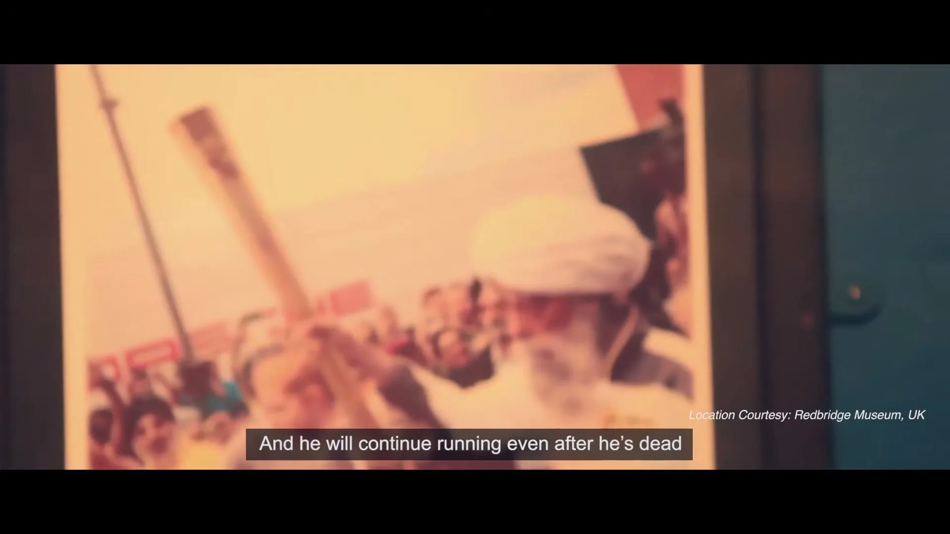 A framed photograph of an elderly Sikh man with a white turban and beard, holding an Olympic torch, is shown in a museum display.