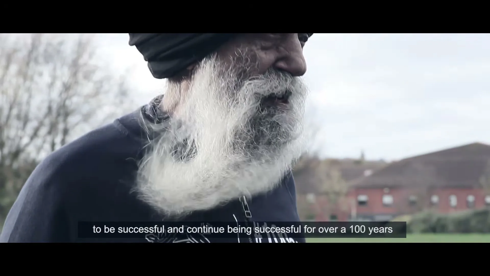 An elderly Sikh man with a long white beard and black turban, wearing a dark blue jacket with a Nestle logo, is seen from the side, talking outdoors.