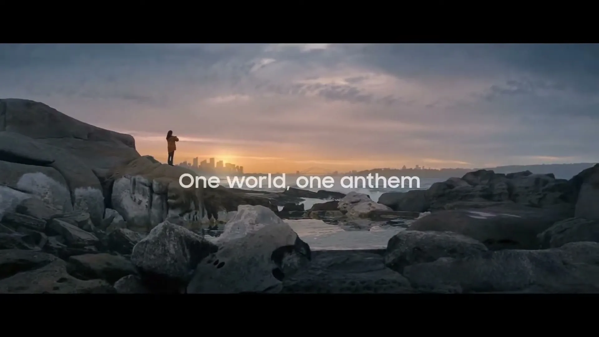 A person stands on a rocky shore, looking out at a city skyline and ocean at sunset, with text appearing on screen.