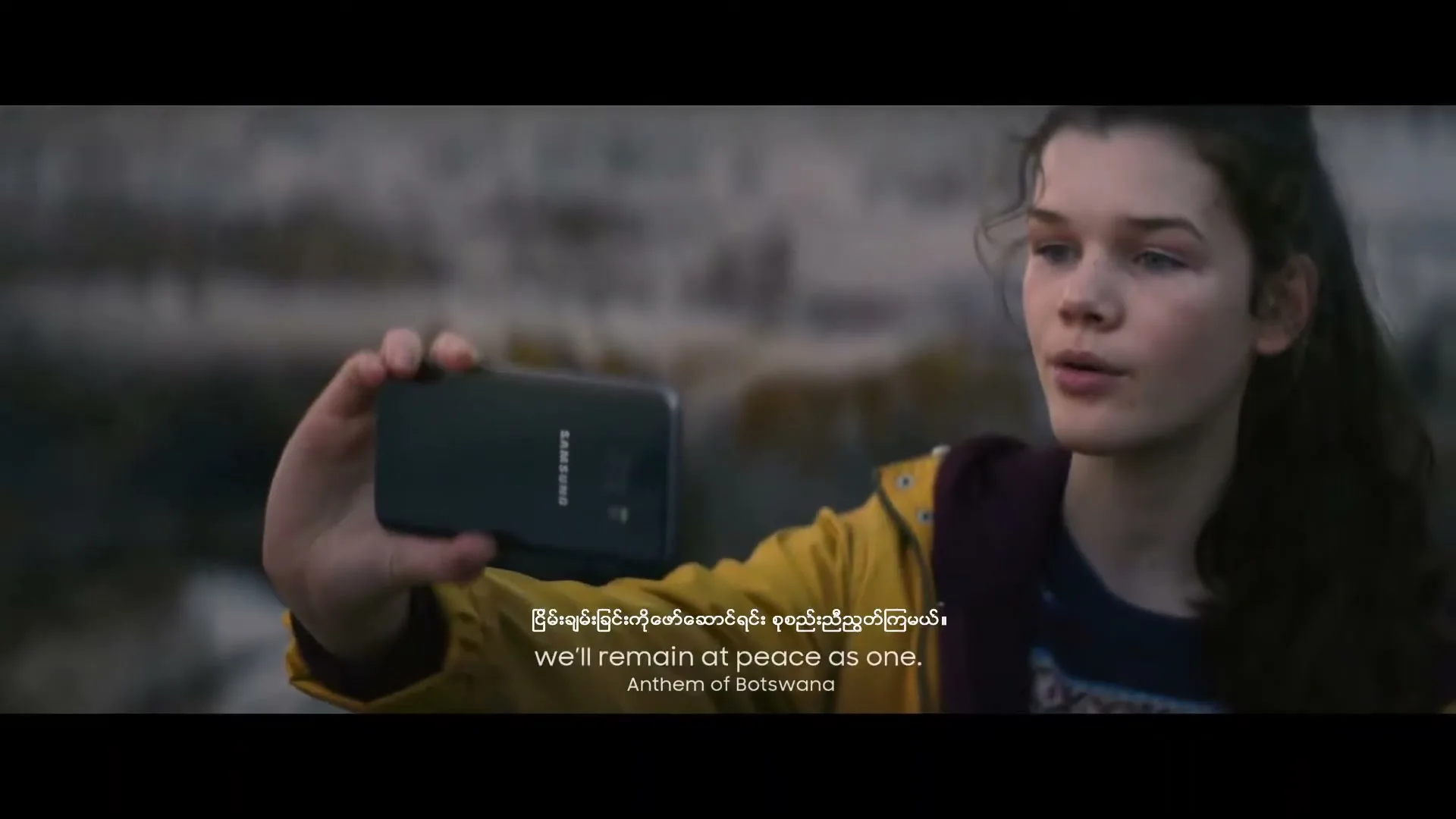 A young girl with long brown hair, wearing a yellow jacket, holds up a black Samsung phone to take a selfie outdoors.