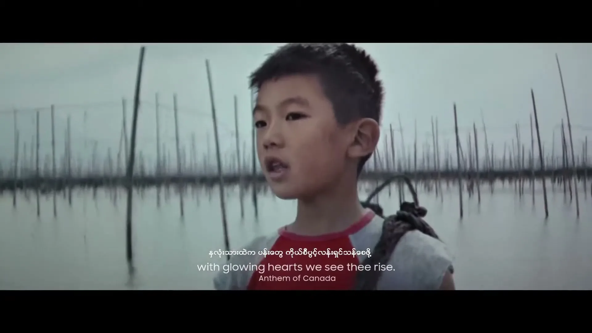 A young Asian boy with short dark hair and a red and grey shirt stands in a shallow body of water with many vertical poles, singing.