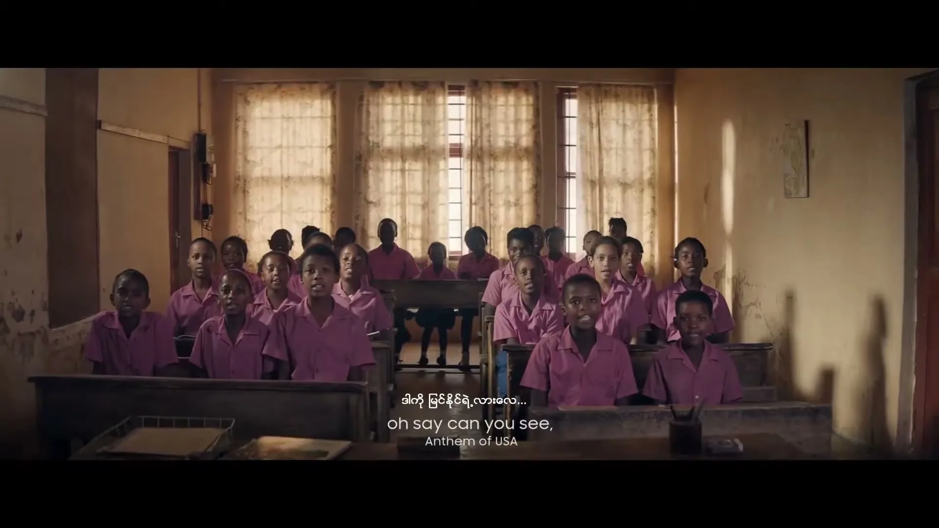 A group of diverse children in pink uniforms sing in a classroom with open windows, their mouths wide open in song.