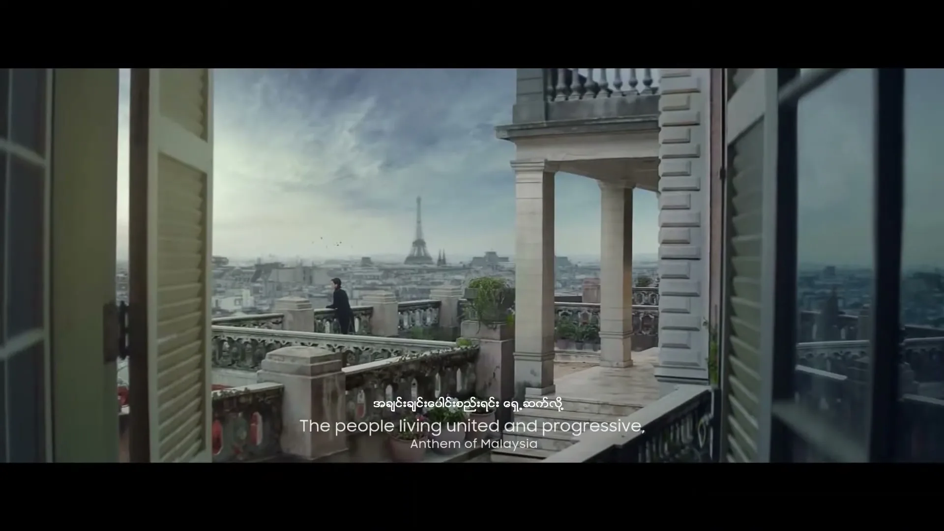 A man in a dark suit stands on a balcony overlooking the Paris skyline with the Eiffel Tower in the distance, seen through open French doors.