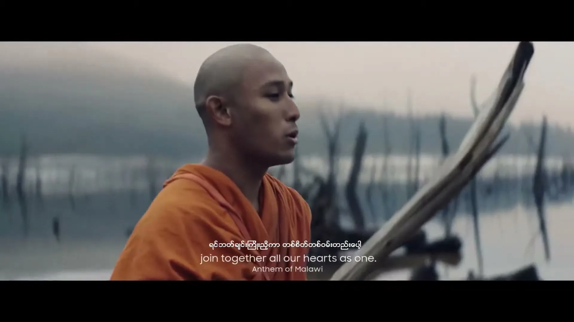 A young, bald monk in an orange robe paddles a wooden boat on a misty lake with submerged trees, singing with a focused expression.