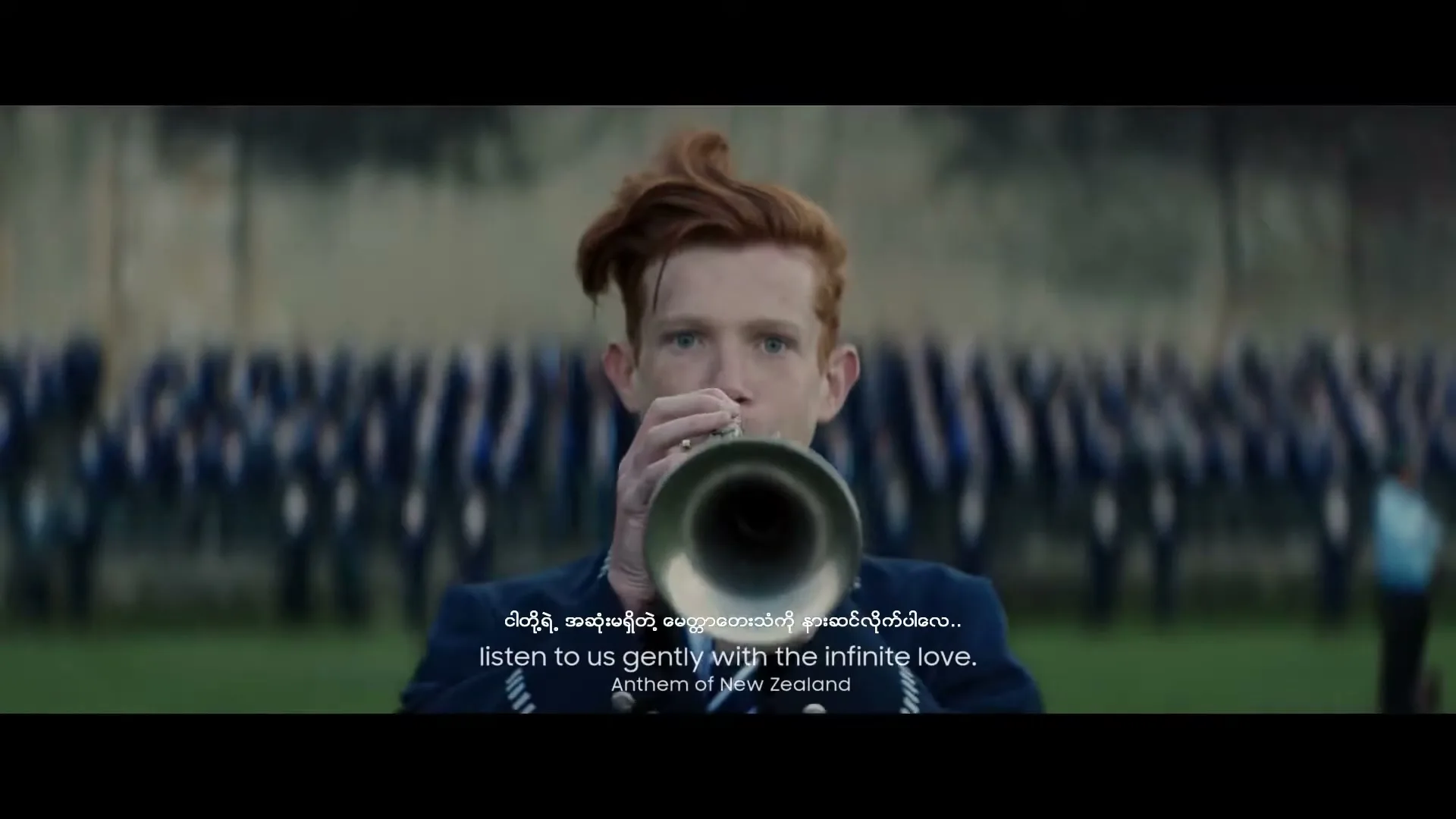 A young man with red hair plays a trumpet in a marching band uniform, with other band members blurred in the background on a grassy field.