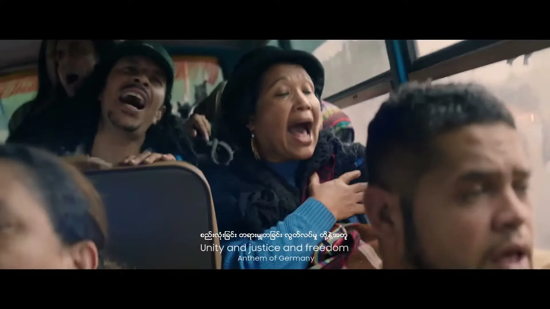 A diverse group of people, including a woman in a black hat and blue sweater, sing passionately on a bus, with text overlaying the scene.