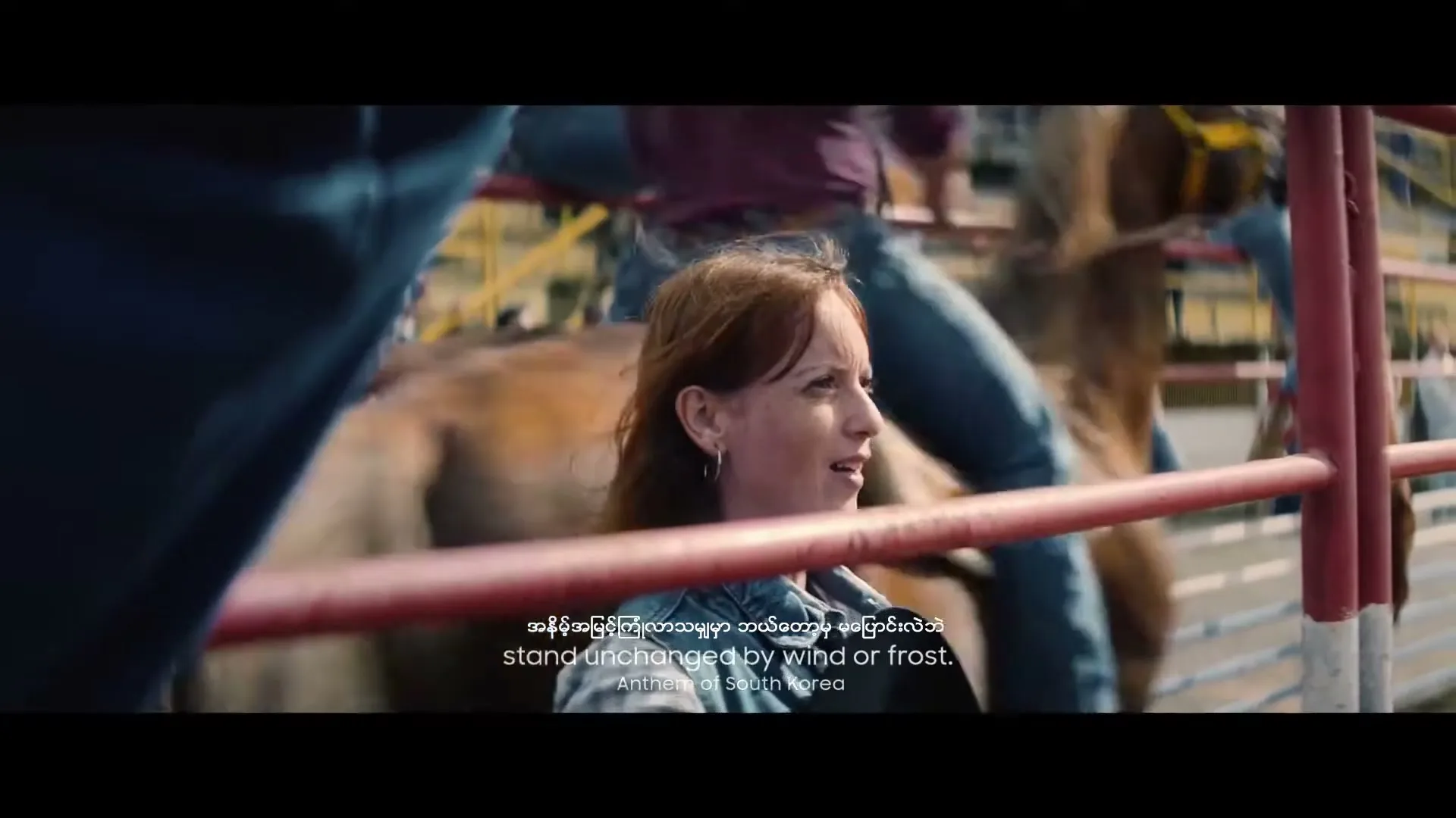 A woman with red hair in a denim jacket watches a rodeo event from the stands, with people and horses in the background.