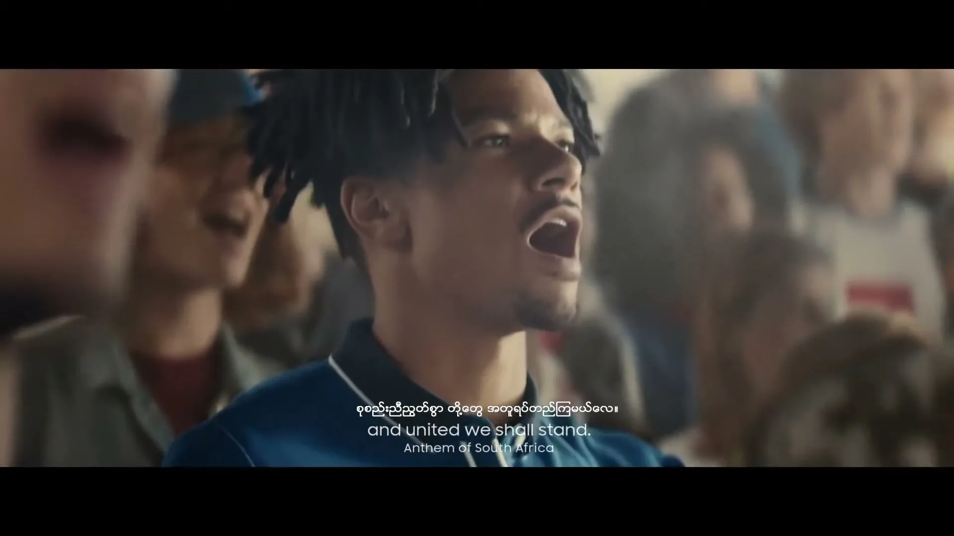 A young man with dreadlocks sings passionately in a crowd, wearing a blue polo shirt, in a stadium.