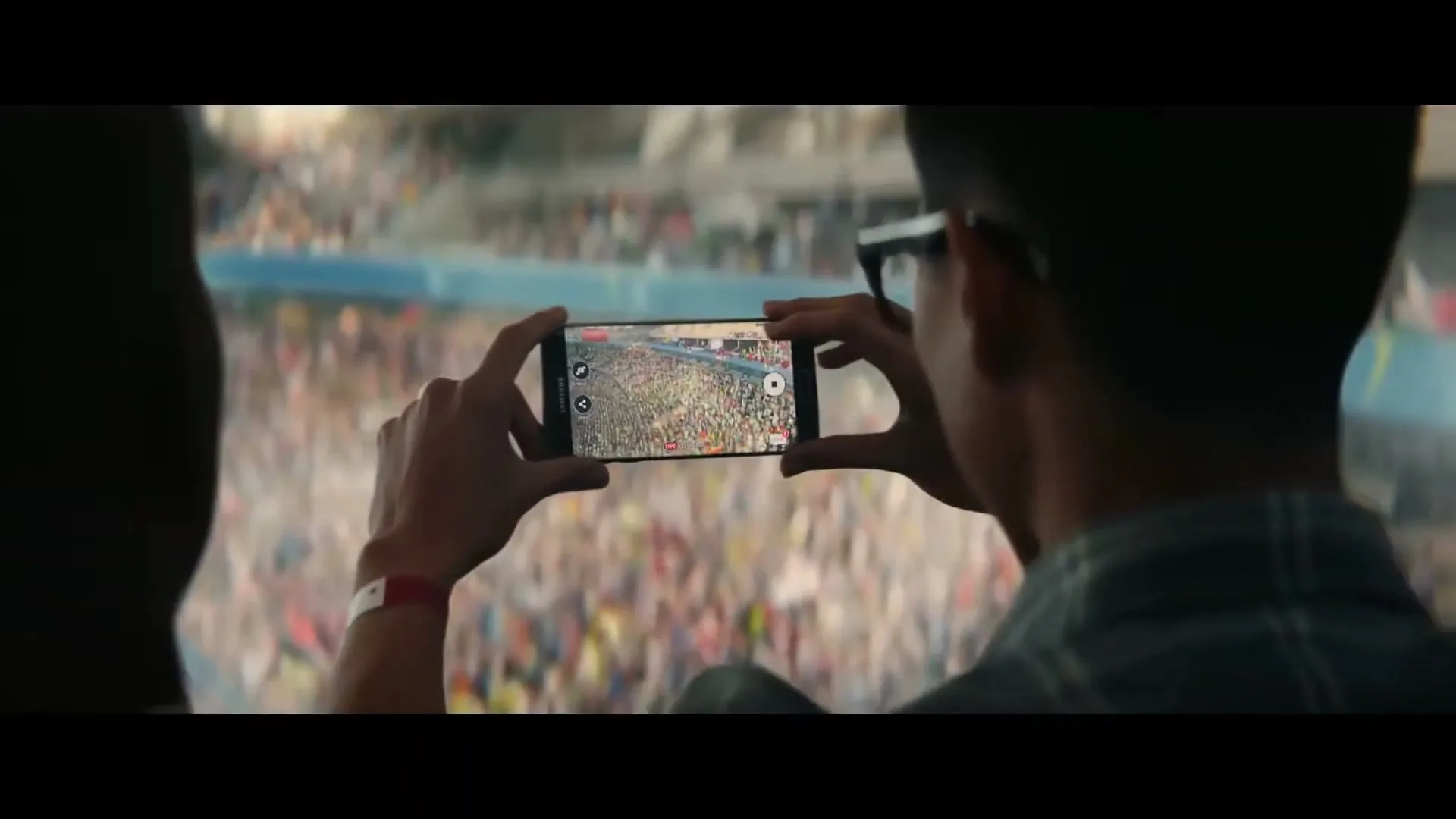 A young man in glasses holds a Samsung phone horizontally, recording a large crowd in a stadium from a high vantage point.