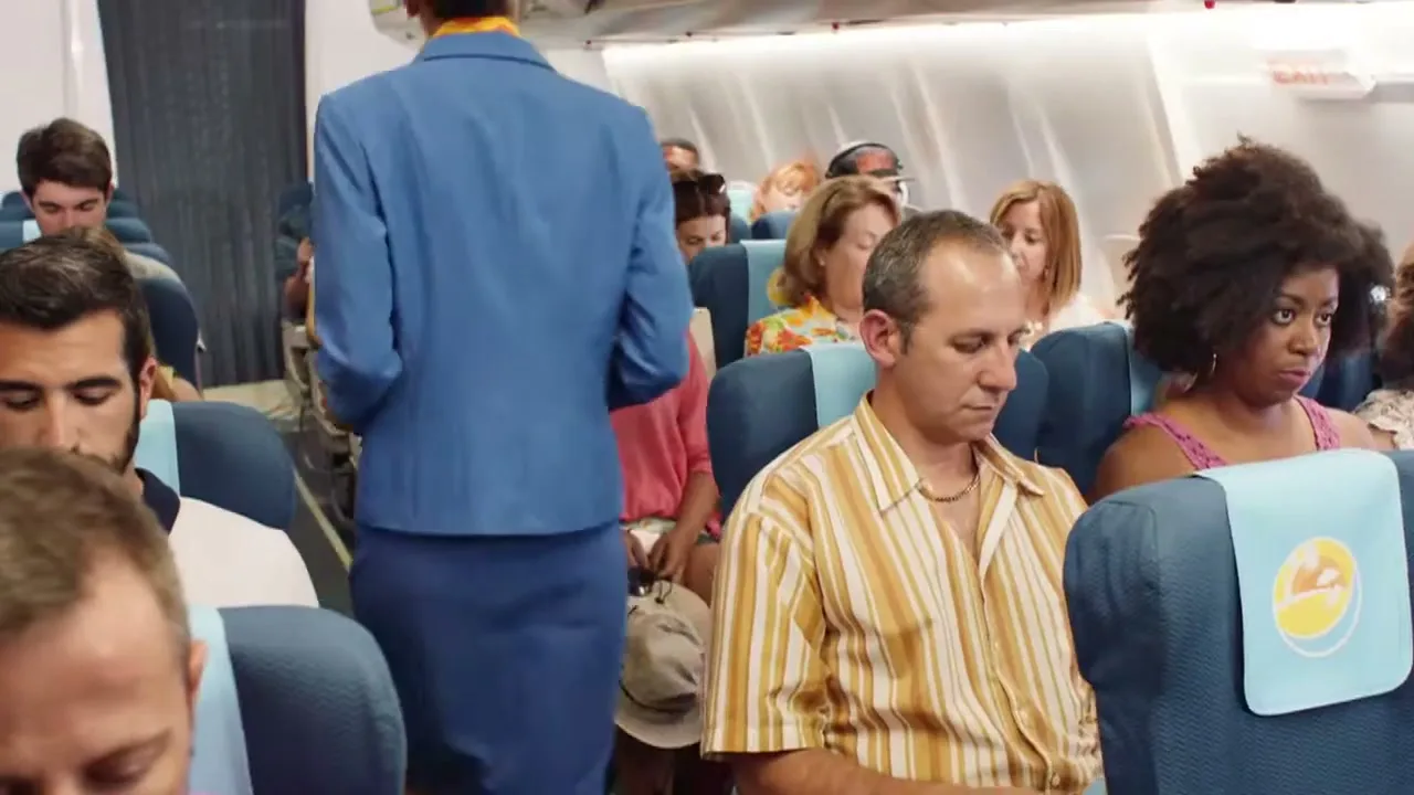 A flight attendant walks down the aisle of an airplane, passing by passengers who look bored or tired.