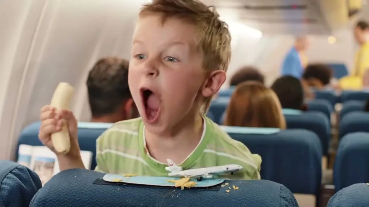 A young boy with blonde hair in a striped t-shirt holds a banana and yells excitedly while playing with a toy airplane on an airplane seat.