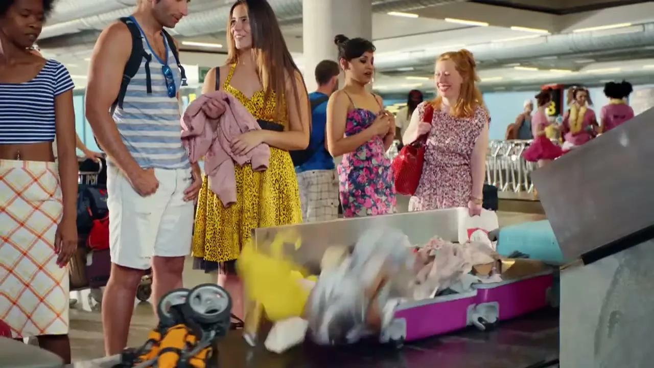 A group of diverse young adults stand at an airport baggage claim, watching as their luggage, including an open suitcase with clothes spilling out, moves along the conveyor belt.