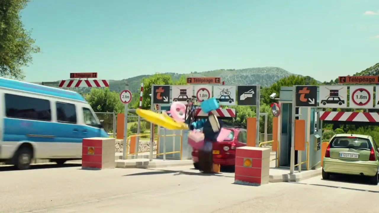 A red car with luggage on its roof crashes into a toll booth, scattering items everywhere, while other cars pass by on a sunny day.