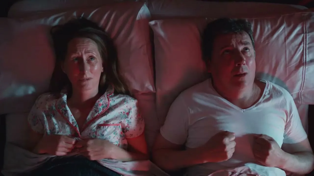 A man and woman lie awake in bed, looking worried and sweating, illuminated by a red glow in a dark bedroom.