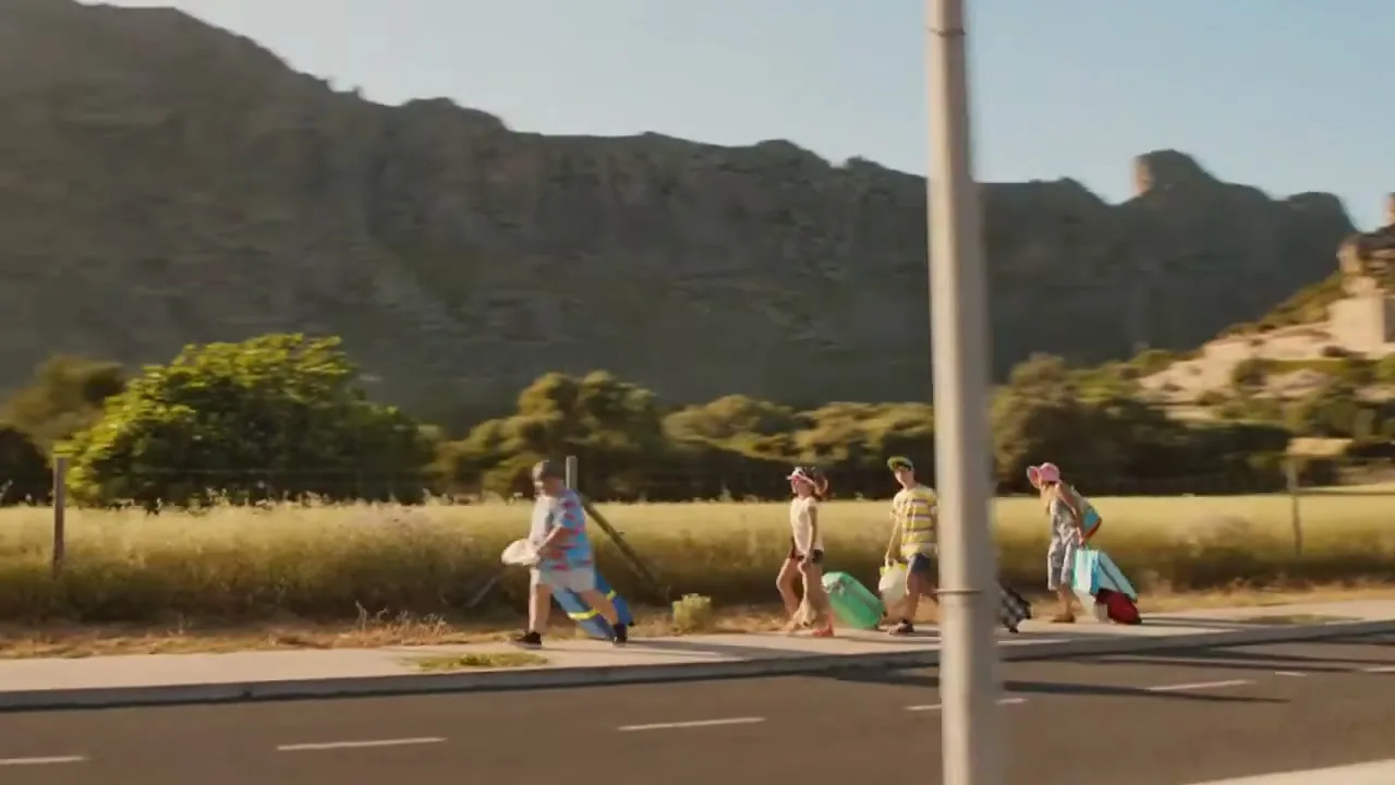 A family of four walks along a road with luggage, with mountains and fields in the background, under a bright sky.