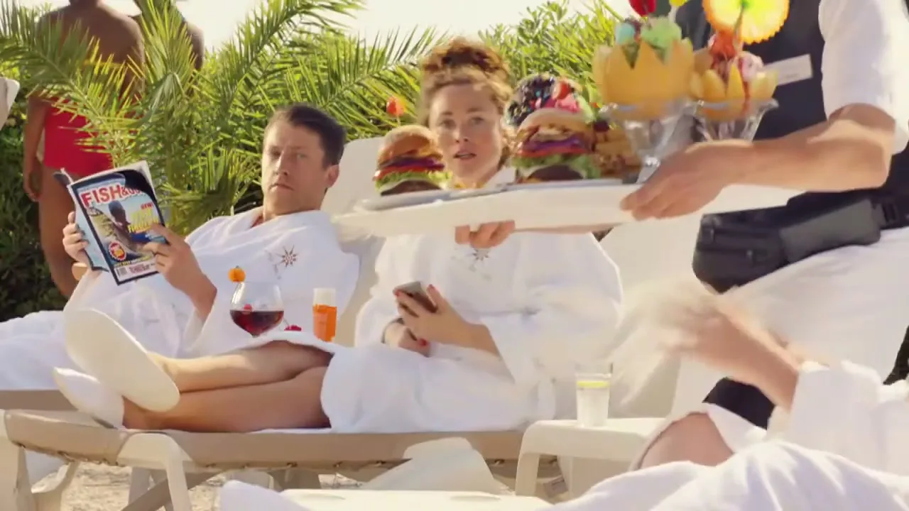 A man and woman in white robes relax on sun loungers by a pool, as a waiter serves them an extravagant tray of food and drinks.