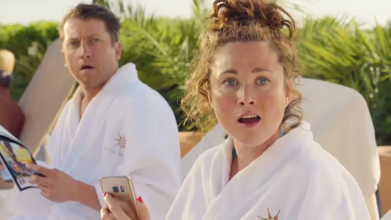A man and a woman in white bathrobes look surprised while relaxing on sun loungers by a pool, with palm trees in the background.