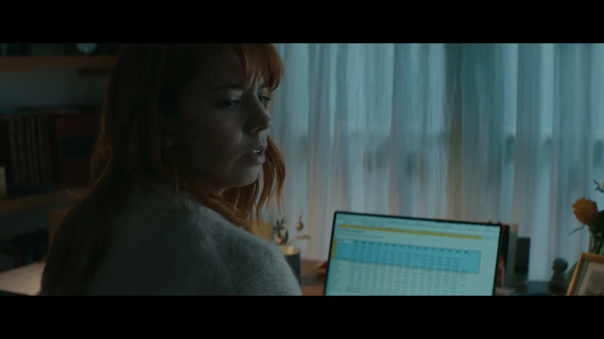 A young woman with red hair, wearing a white sweater, works on her laptop at a desk in a dimly lit room, then turns to look over her shoulder.