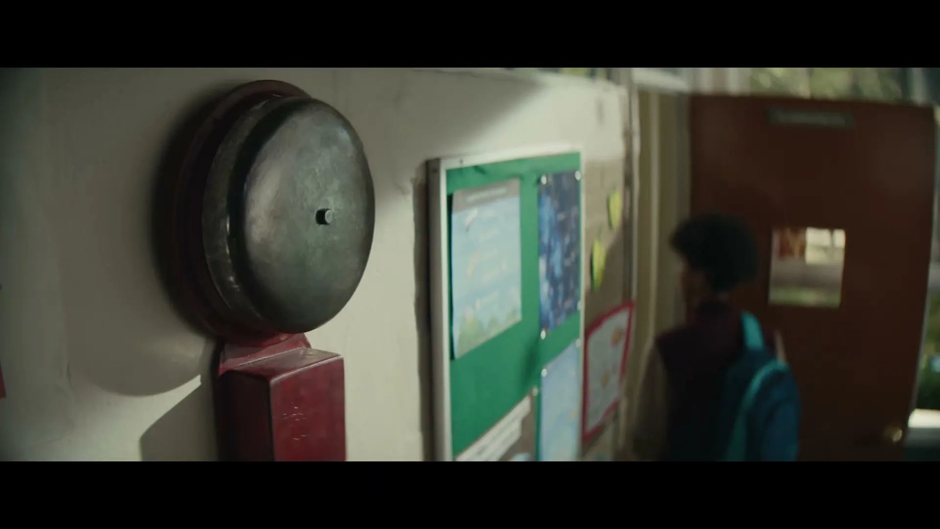 A school bell is mounted on a wall next to a bulletin board, as a student walks past a classroom door in a school hallway.