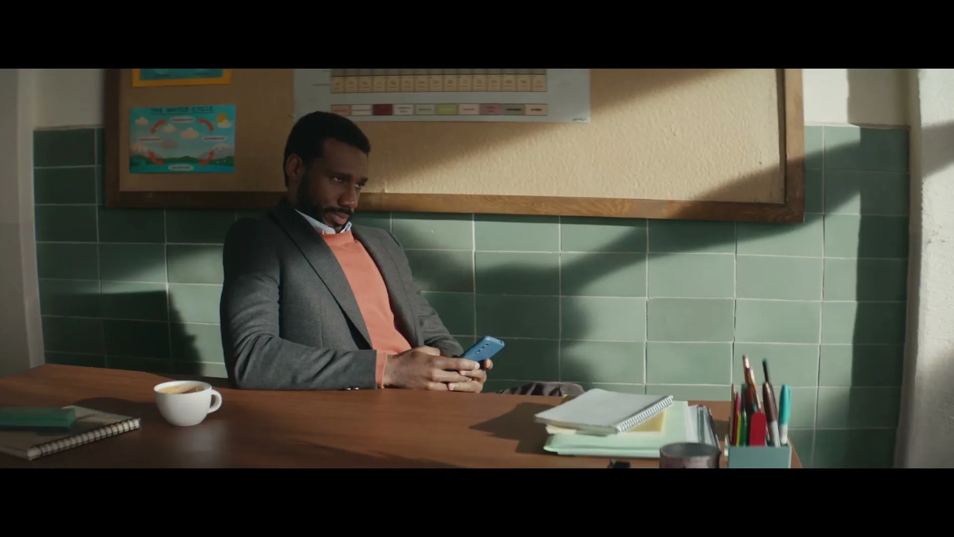 A Black man in his 30s, wearing a suit jacket and orange sweater, sits at a desk in a classroom, looking at his phone.