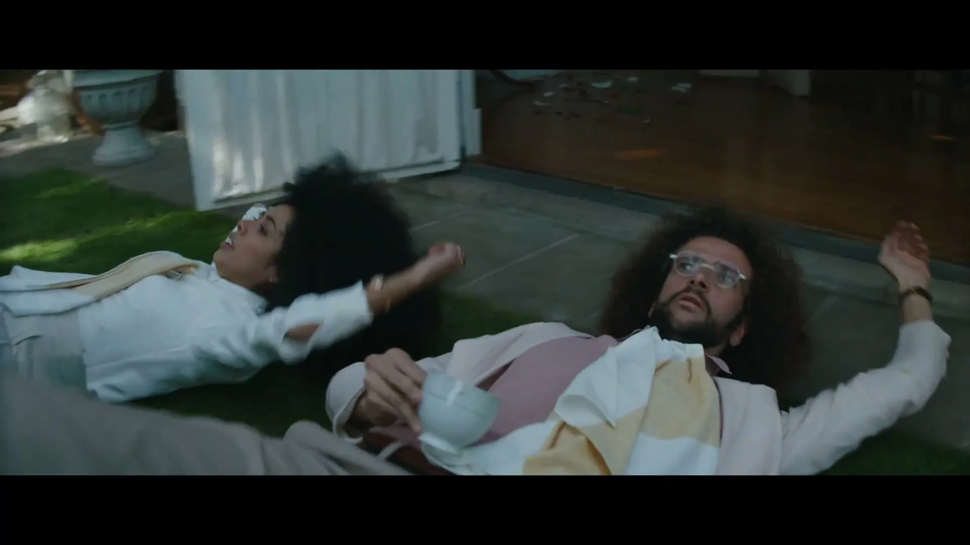 A man and a woman with curly hair fall backward onto a green lawn outside a house, looking surprised.