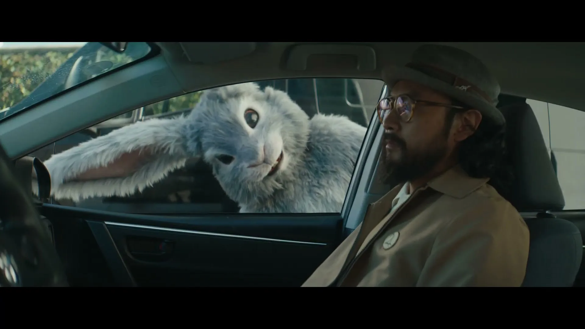 A man in a car looks at a giant rabbit with long ears and a creepy smile, peering through the car window.
