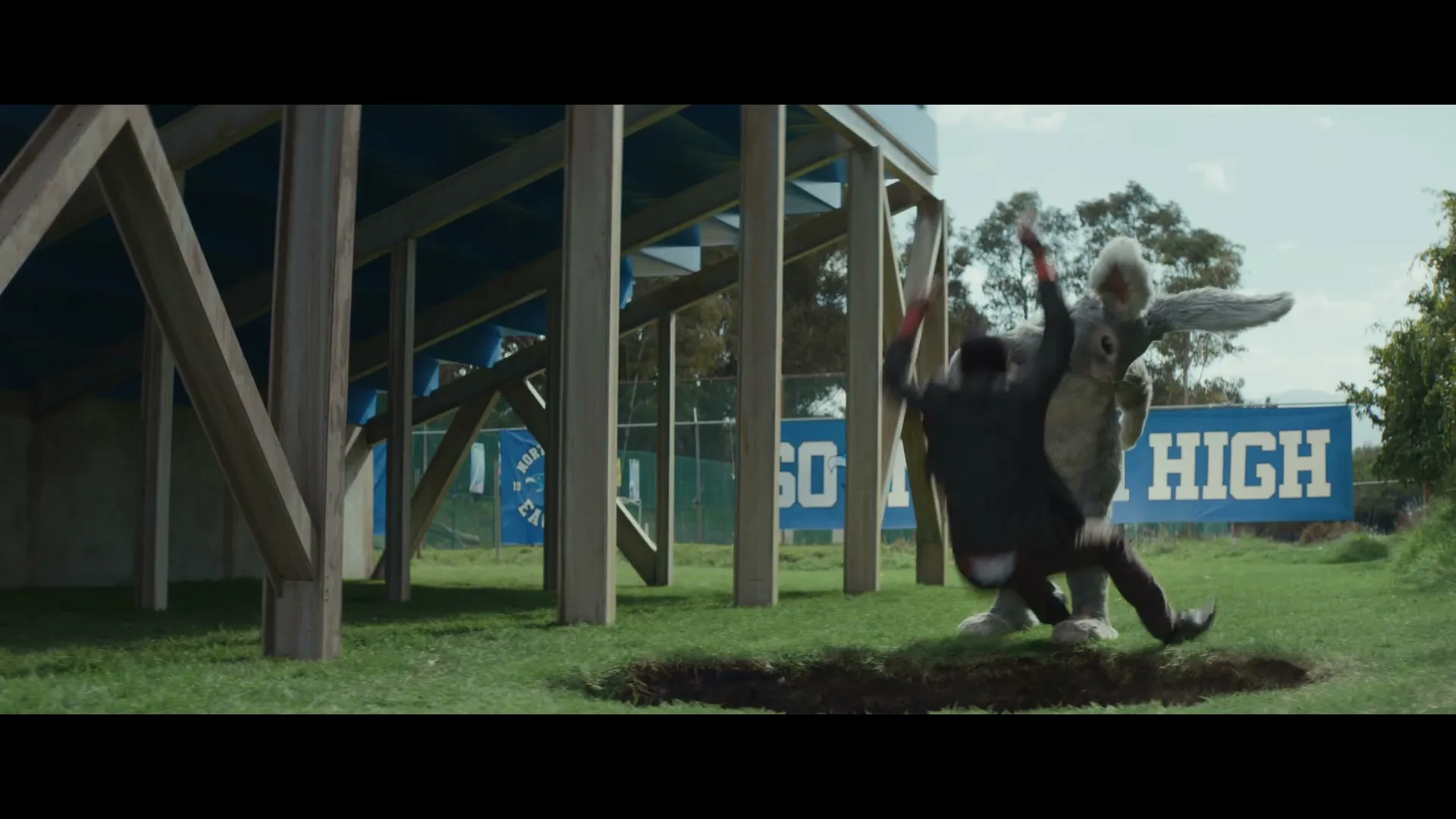A man in a suit is thrown off a giant rabbit costume near a hole in the ground next to bleachers on a sunny day.