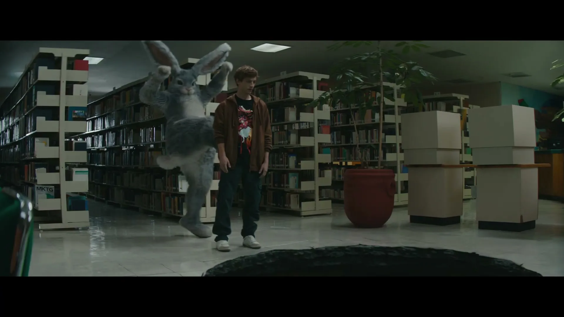 A young man in a library is surprised by a large, grey rabbit-like creature that emerges from behind a bookshelf and pushes him into a dark hole on the floor.