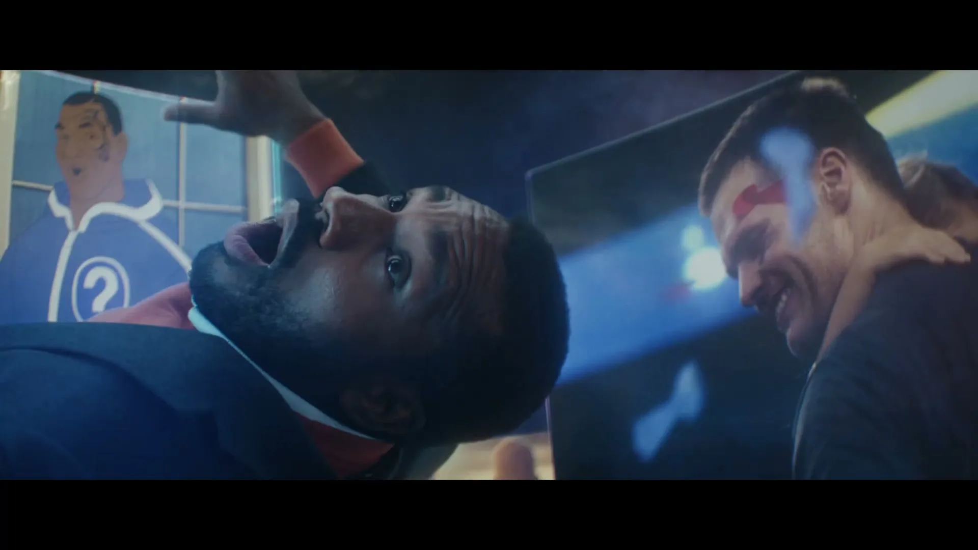A man in a suit is screaming while lying down, with another man in a black shirt smiling next to him, in a dark, blue-lit room.