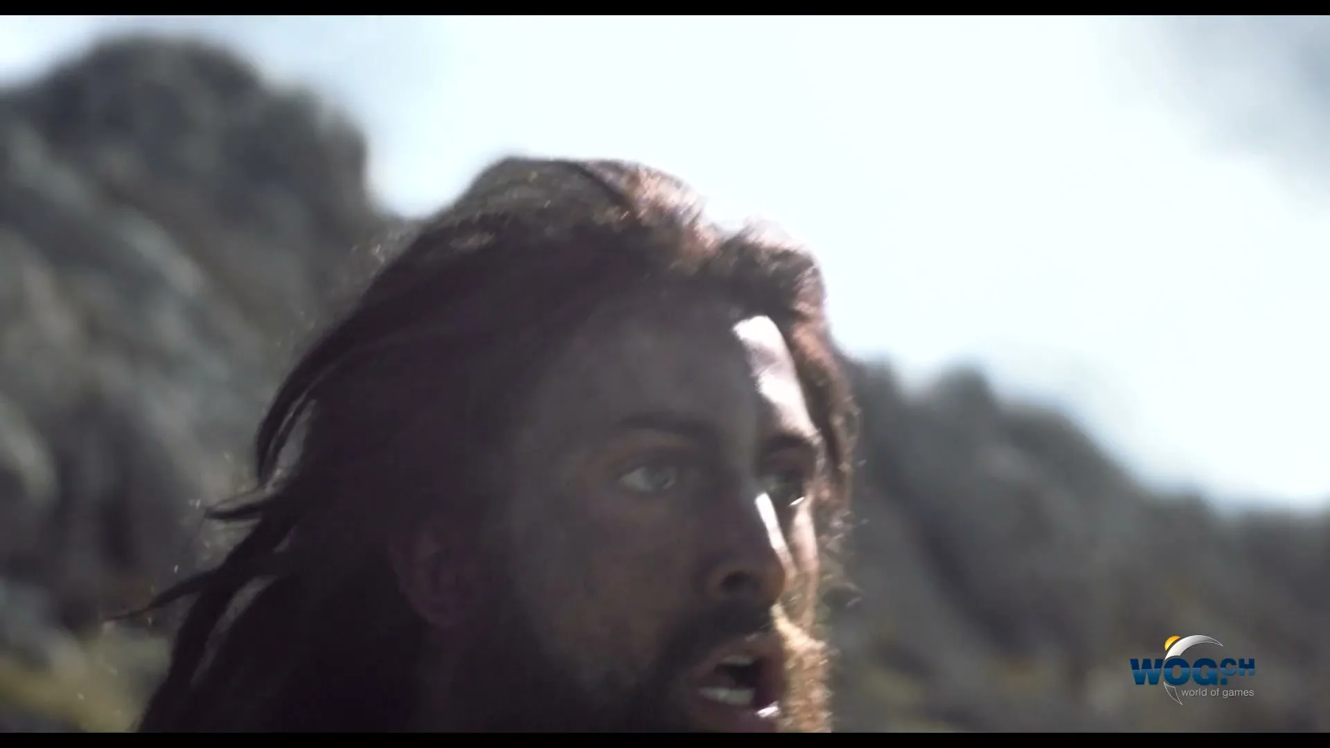 A man with long dark hair and a beard, wearing primitive clothing, looks up with a worried expression in a rocky, mountainous landscape.