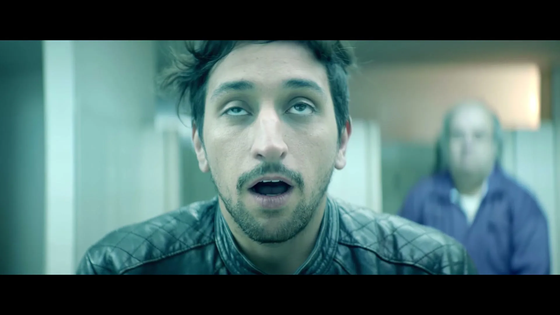 A man with messy hair and a leather jacket looks up with wide eyes, while an older man in a blue shirt is out of focus in the background.
