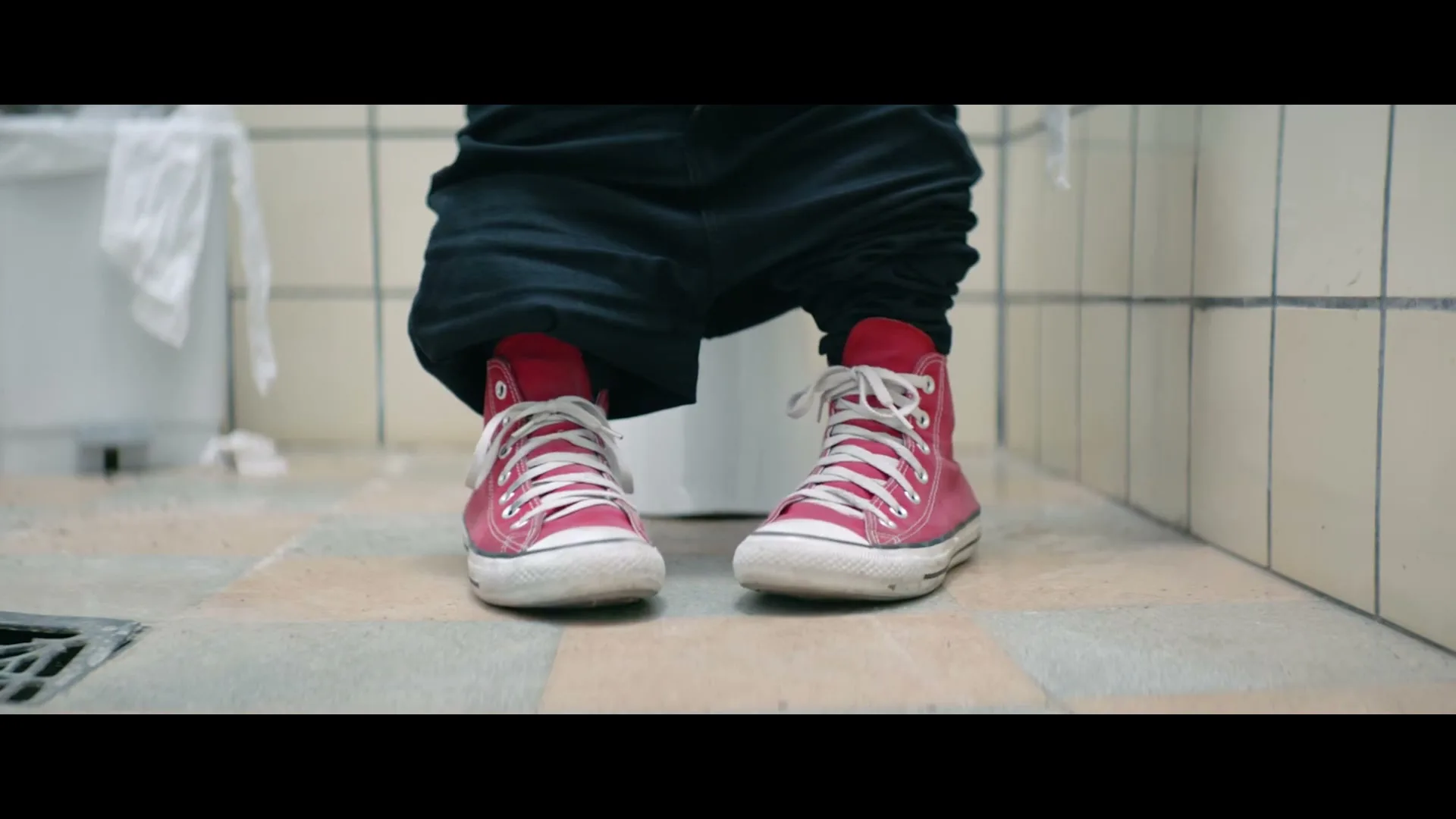 A person wearing black pants and red high-top sneakers sits on a toilet in a public restroom, with their pants around their ankles.