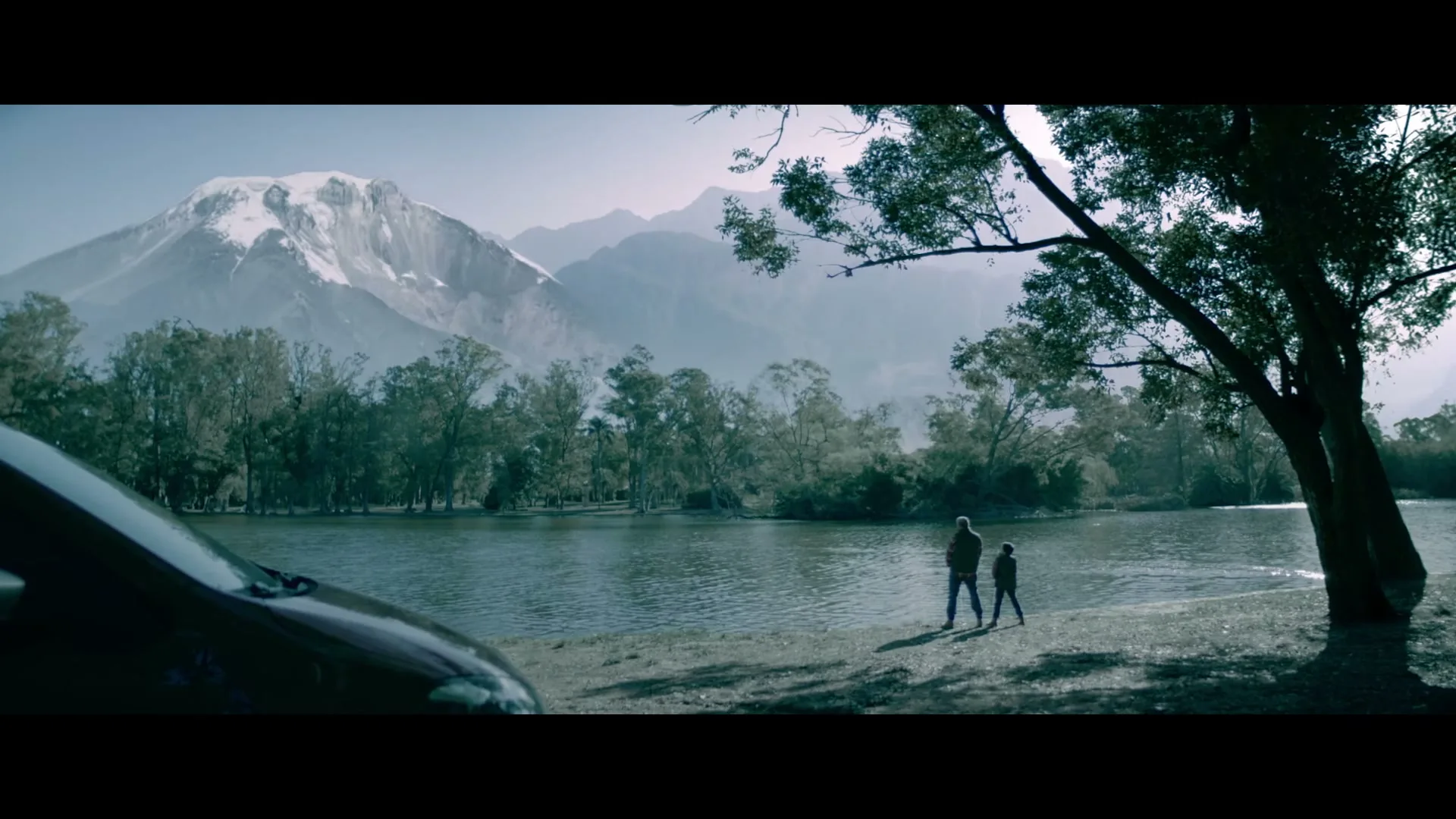 A father and son stand by a lake with a snow-capped mountain in the background, next to a car.