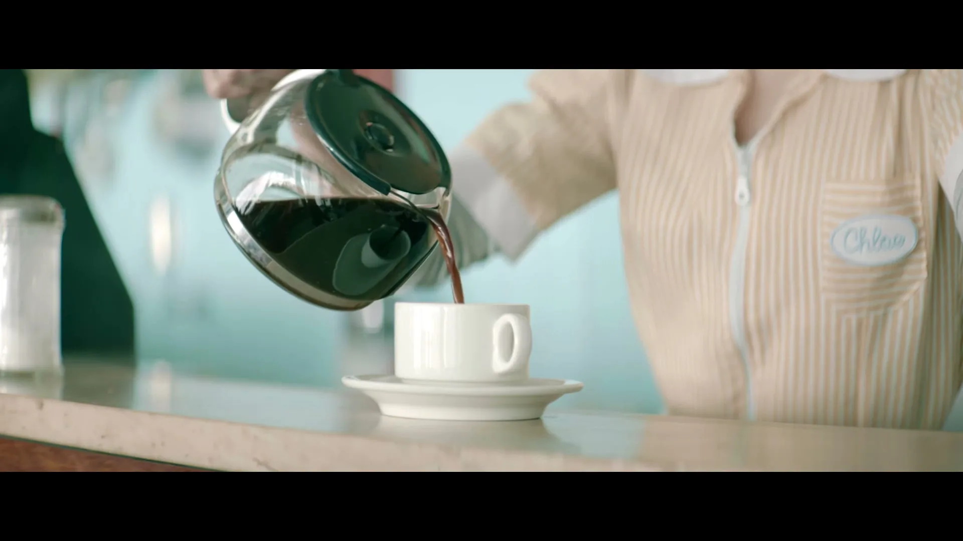 A female diner waitress in a striped uniform pours coffee from a glass pot into a white cup on a counter.