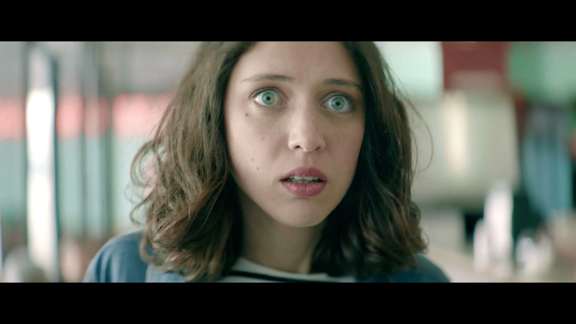 A young woman with wide, surprised eyes and curly brown hair looks directly at the camera, then glances to the side in a brightly lit interior.