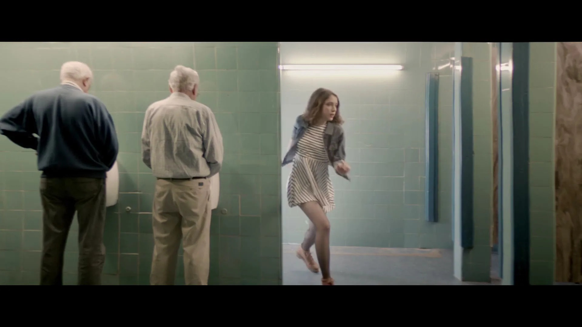 Two older men stand at urinals in a public restroom while a young woman runs past the entrance, looking distressed.