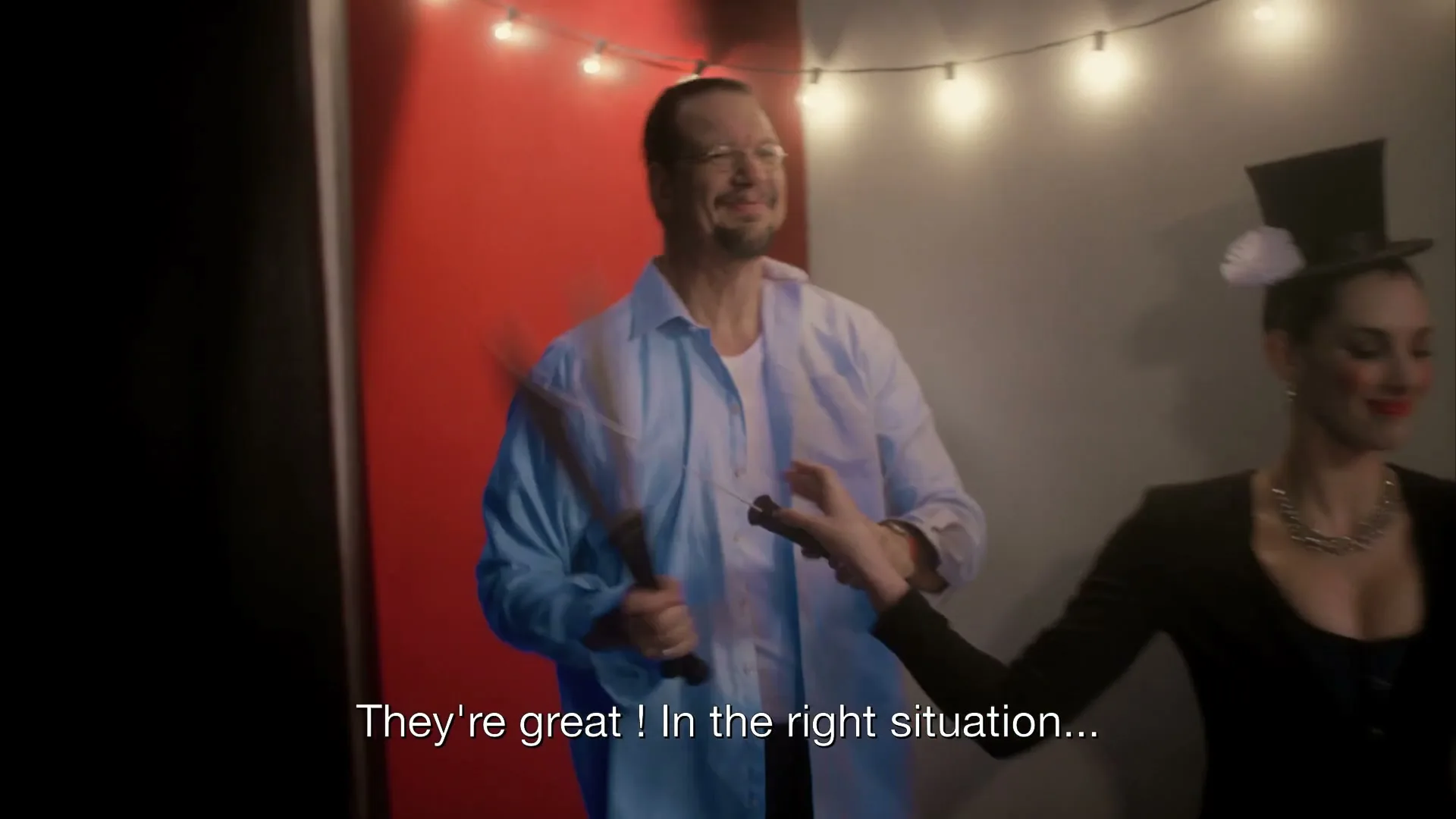 A man in a light blue shirt and glasses juggles knives while a woman in a top hat stands next to him, in a room with red and white walls and string lights.