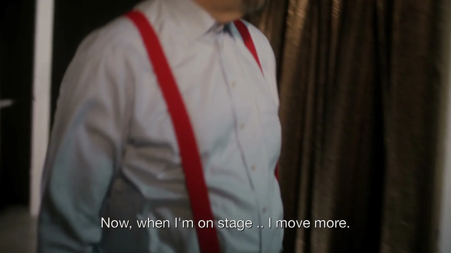 Penn Jillette, a man in his 60s, wearing a white shirt and red suspenders, walks past a woman in a costume, then steps onto a grid of scales on a dark stage.