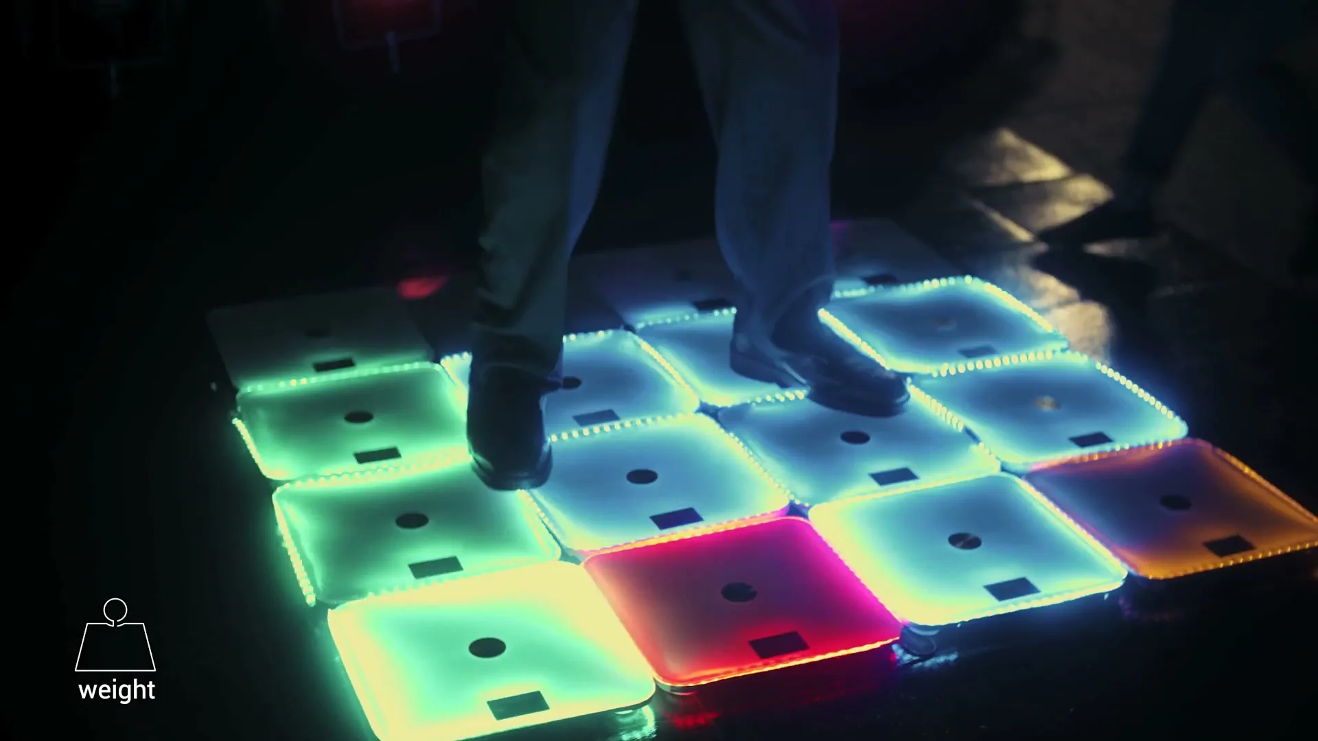 A person's legs and feet are seen from the knees down, stepping on a grid of glowing, colorful smart scales in a dark room.