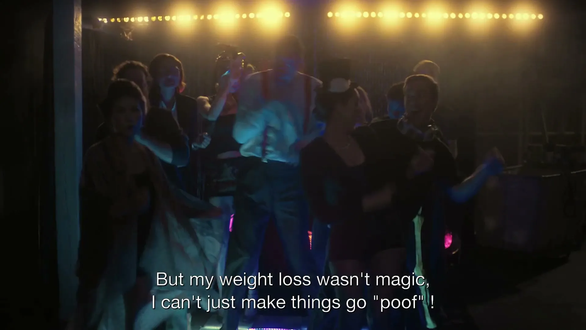 A man in a white shirt and red suspenders dances on a light-up floor with other people in a dark room with bright lights in the background.
