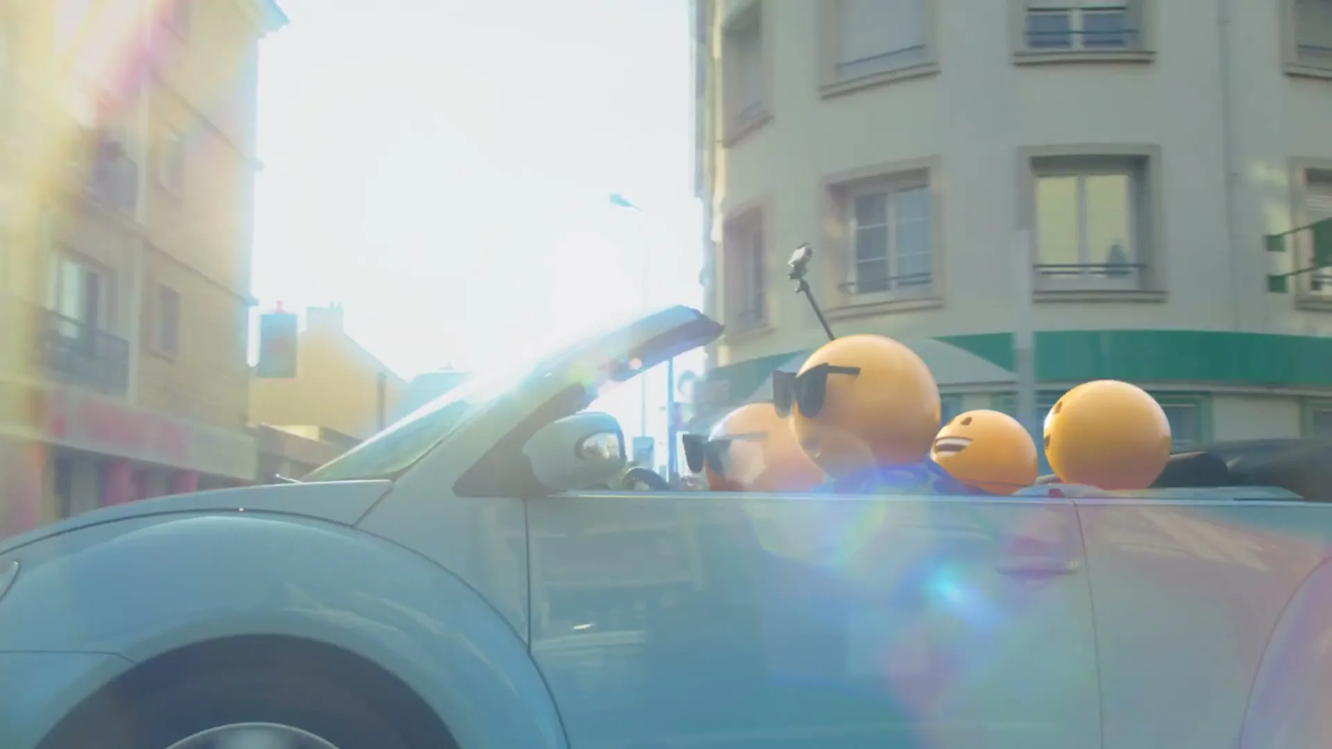 Four emoji characters, one wearing sunglasses, ride in a light blue convertible car down a city street on a sunny day.