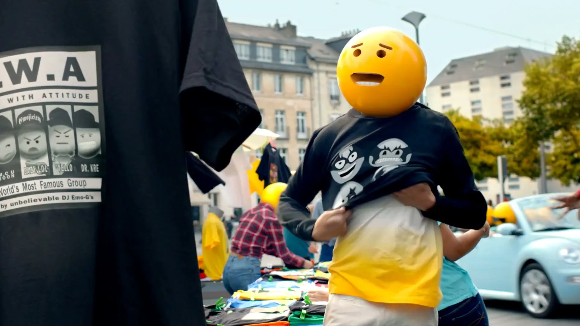 A person with an emoji head tries on a black sweatshirt with emoji faces, standing at an outdoor market stall with an "E.W.A." t-shirt in the foreground.