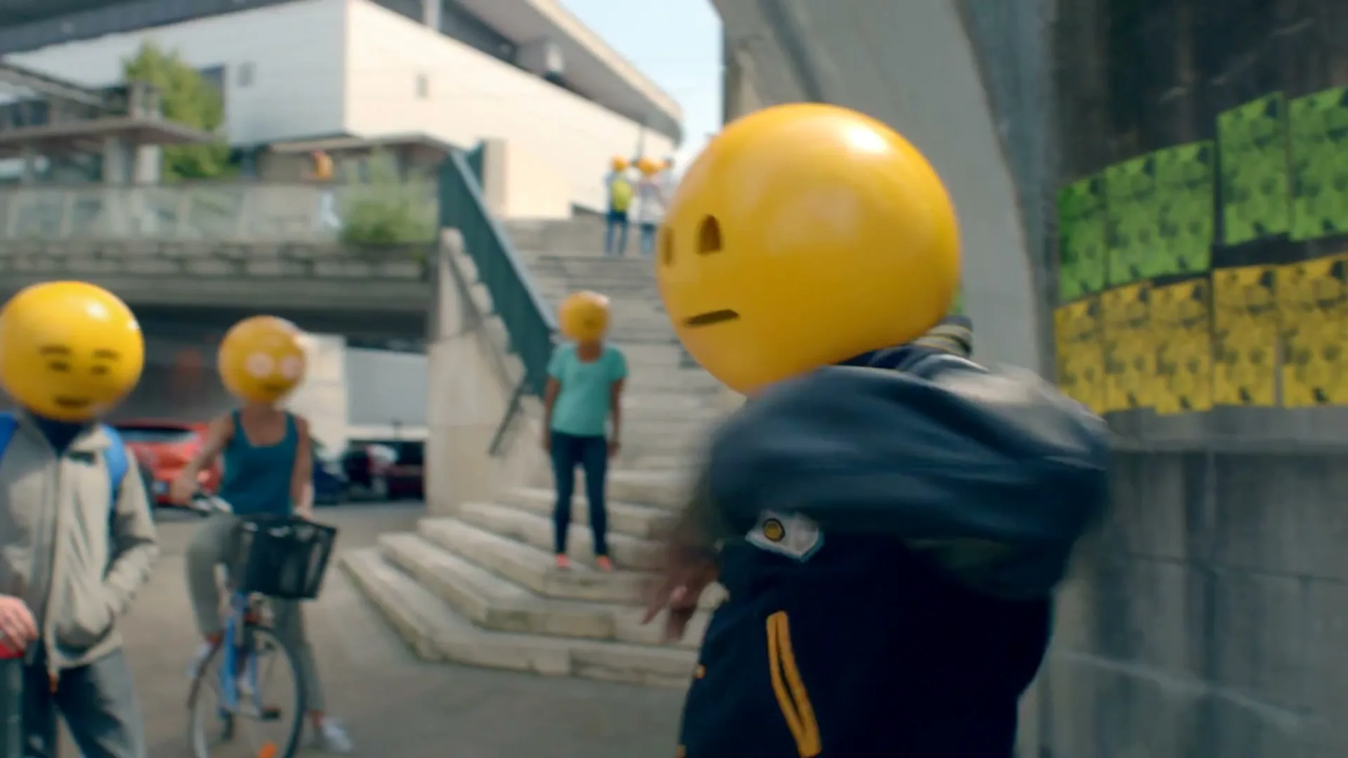 A person in a varsity jacket with a neutral emoji head walks past a wall with posters, as other emoji-headed people are seen in the background on stairs and a bicycle.