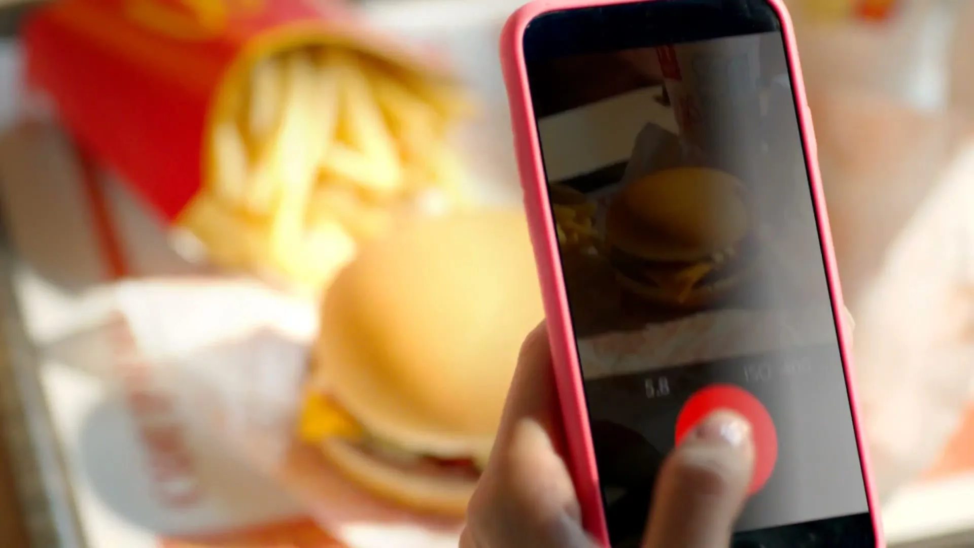 A person holds a pink smartphone, taking a picture of a McDonald's cheeseburger and fries on a tray, with a cartoon burger appearing on screen.