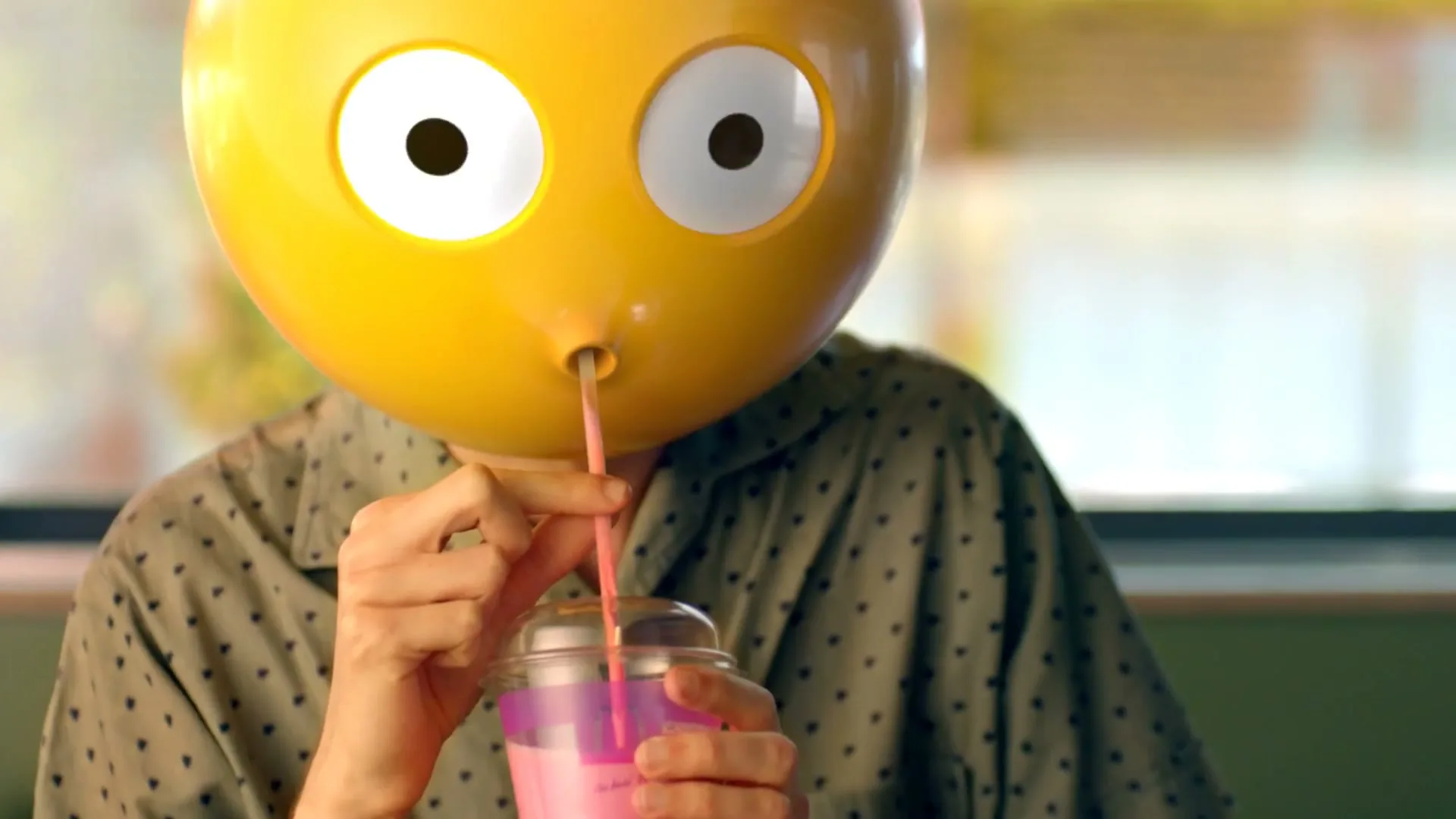A person wearing a yellow emoji mask with wide eyes sips a pink drink through a straw, holding the cup with both hands in an interior setting.