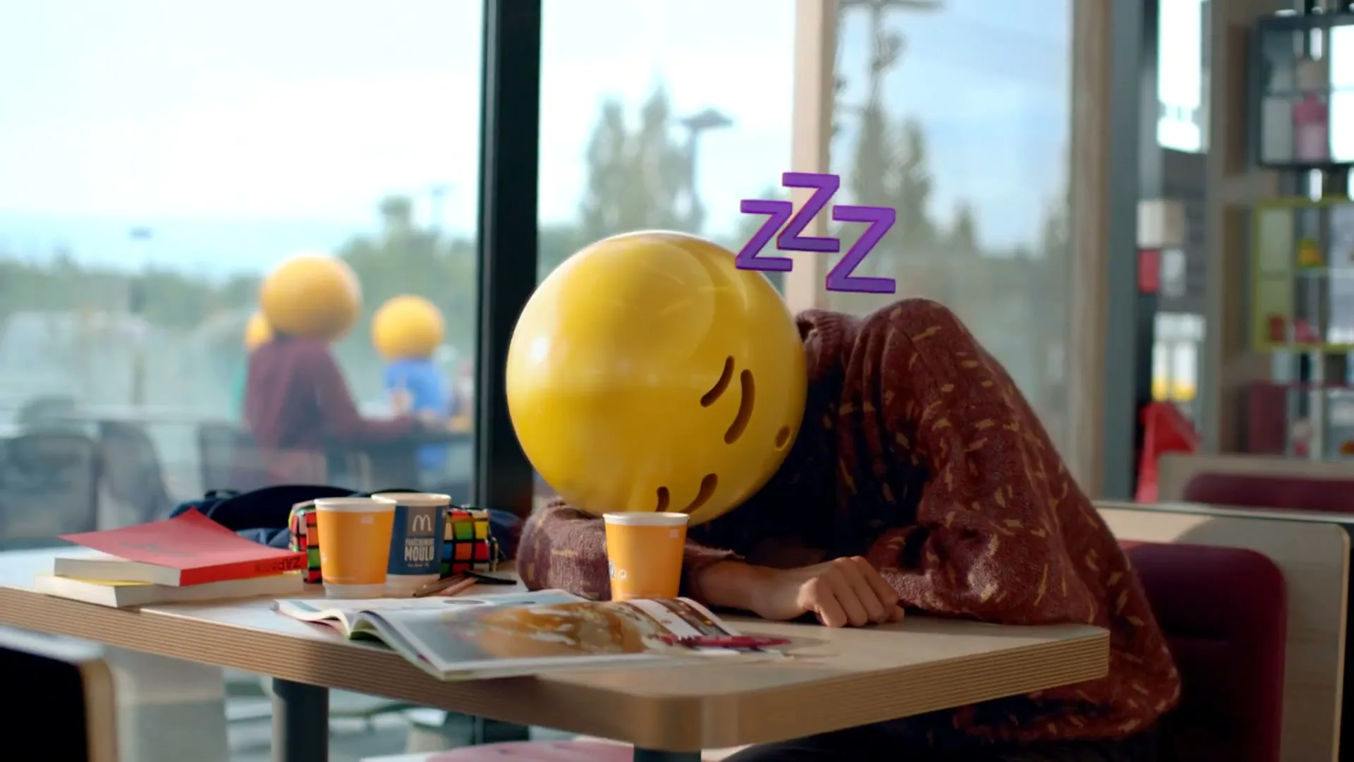 A person wearing a yellow emoji mask with a surprised expression sits at a table in a McDonald's, waking up from a nap.