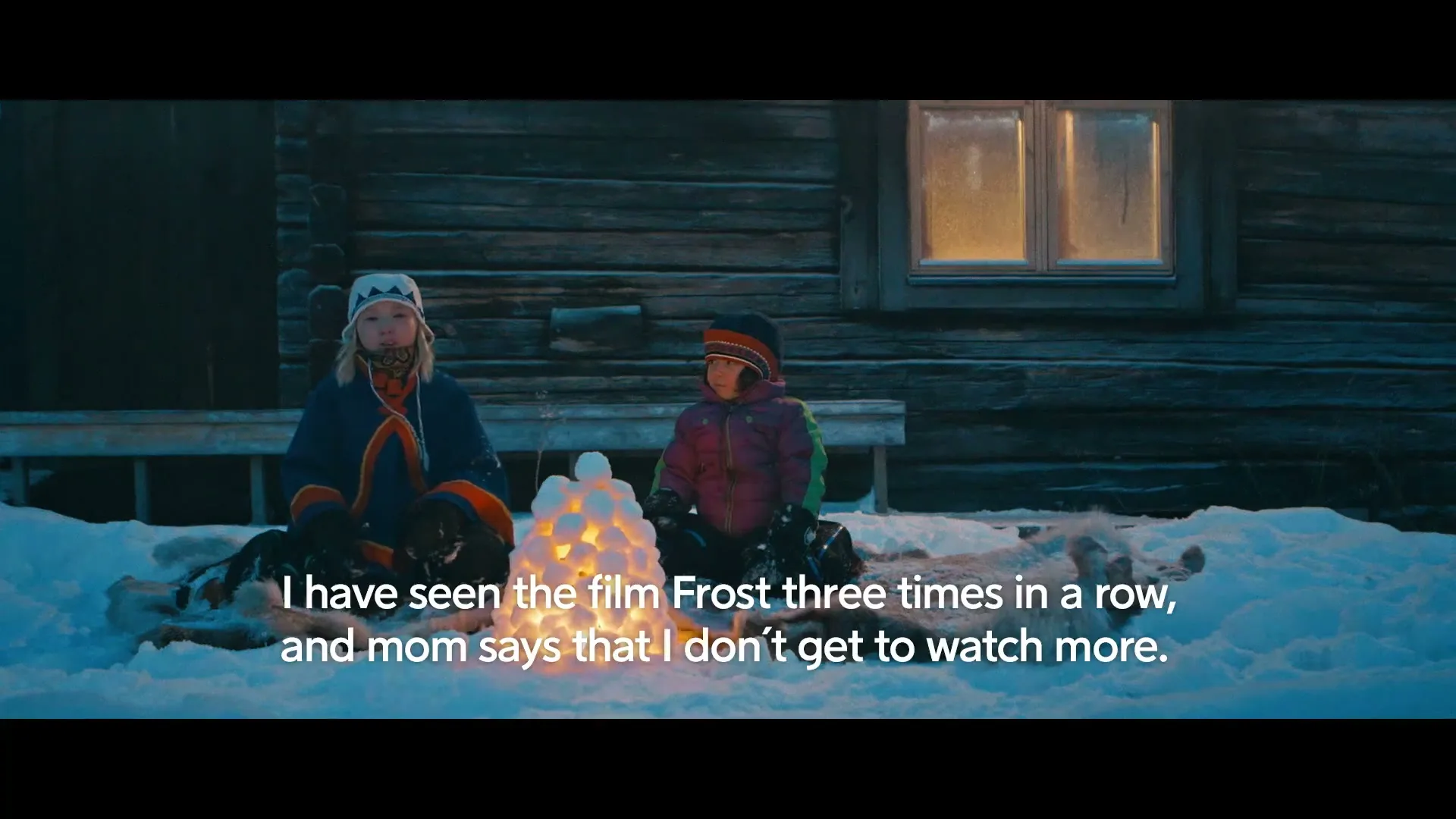 Two children sit in the snow outside a wooden cabin at night, next to a glowing snow sculpture.