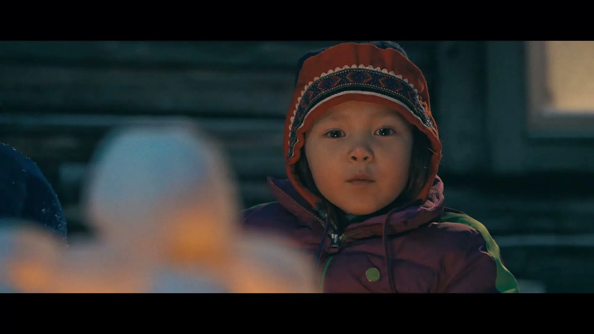 A young child in a winter hat and jacket looks intently at something out of frame, with a blurred foreground element.